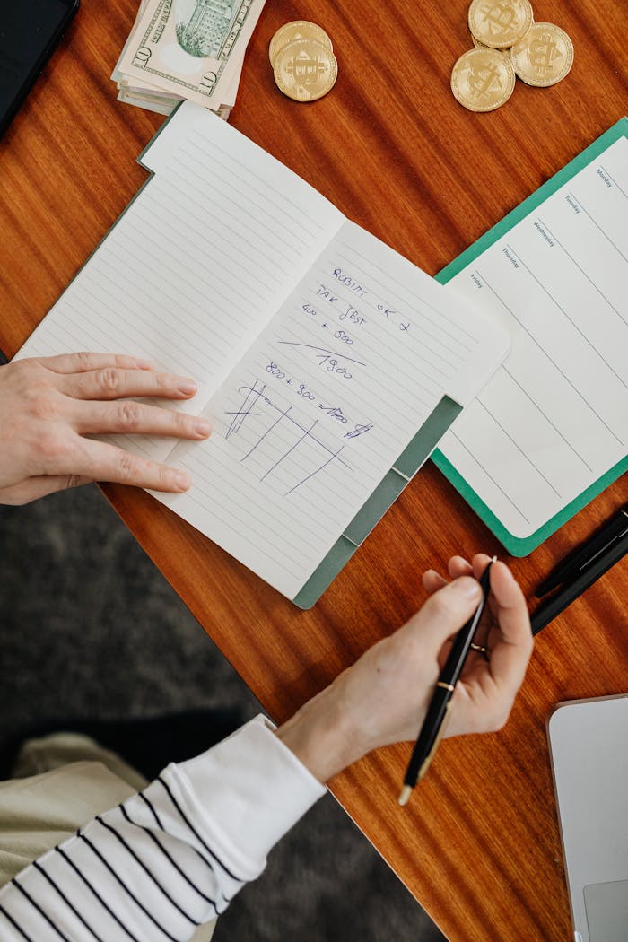 Hands holding pen and writing financial calculations in notebook with cash and coins on wooden desk.